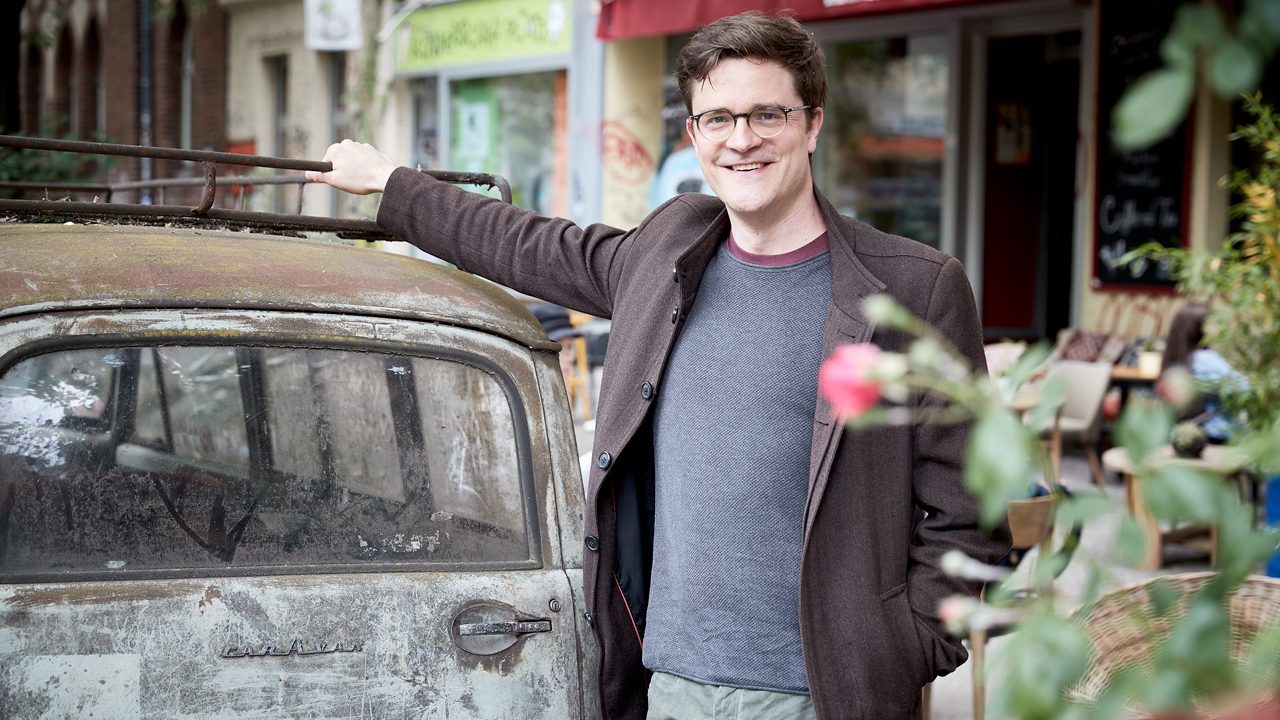 Bodo Wartke lehnt sich an ein verostetes Auto vor dem Café Zazza. Im Vordergrund ein Auto.