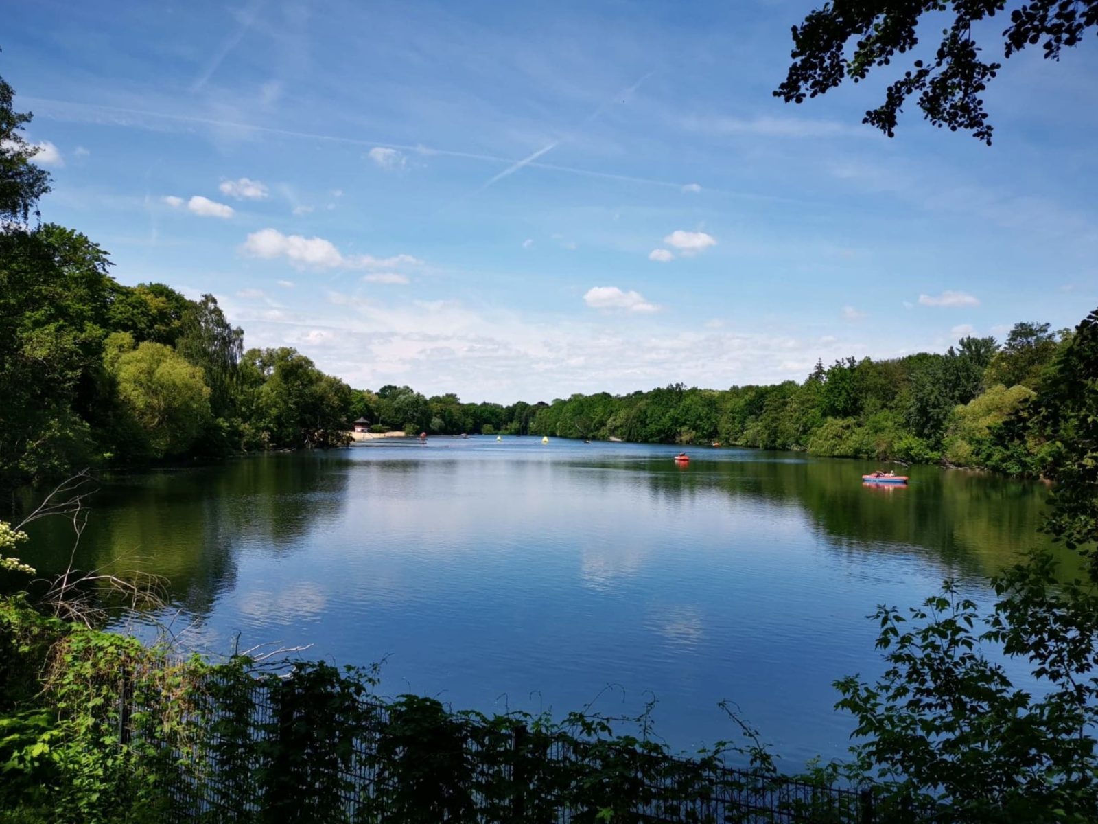 Von BÃ¤umen umstandener See mit zwei Booten, darÃ¼ber blauer Himmel, im Vordergrund Zaun