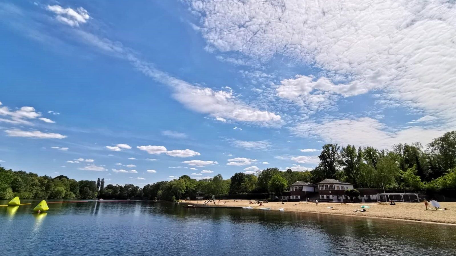 Blick auf den Plötzensee, rechts der Strand und Gebäude des Strandbads, um den See Bäume, blauer Himmel mit Wolken